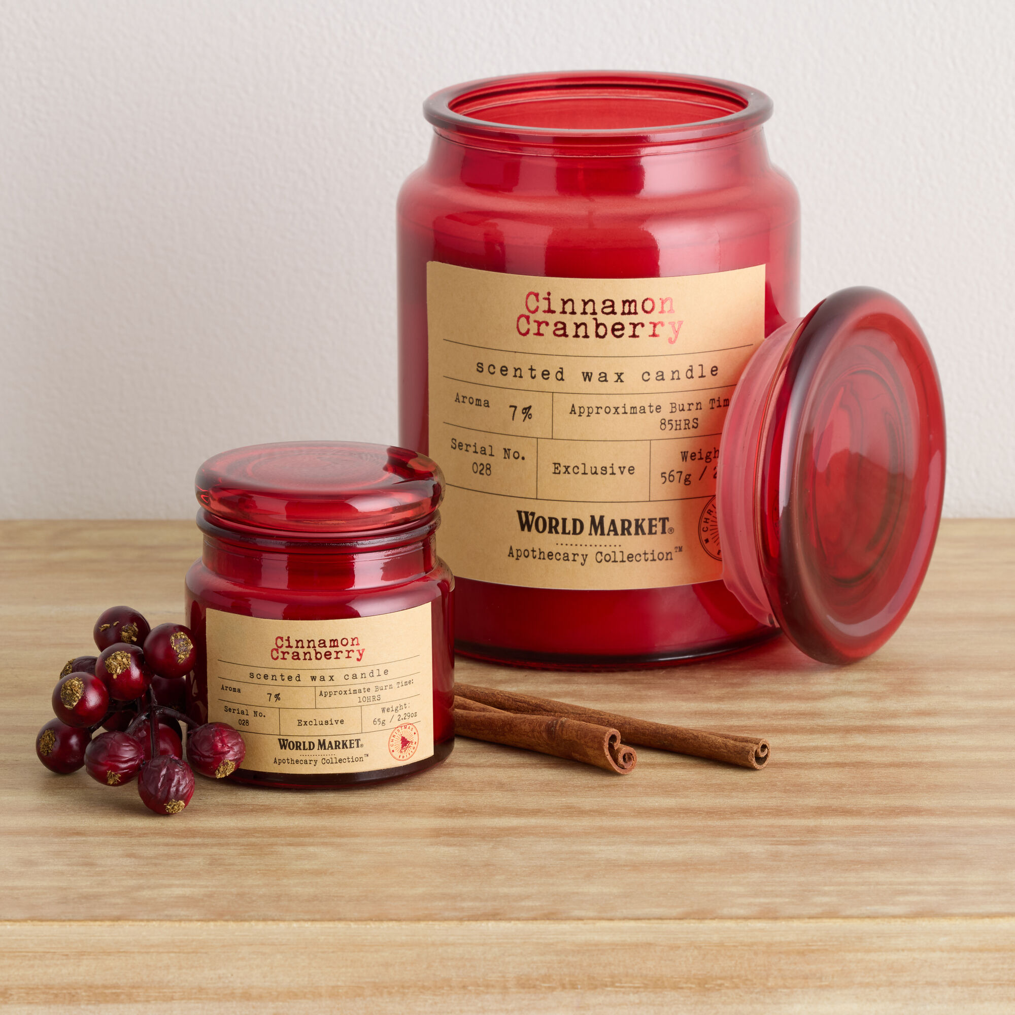 Two red jar candles on a table next to a bunch of cranberries and two cinnamon sticks